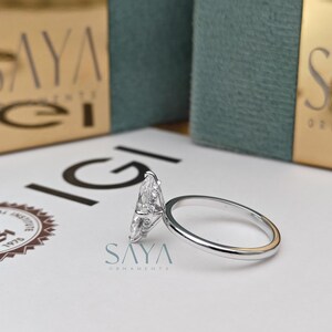 Puede incluir: Un anillo de plata con un diamante talla marquesa, sobre una superficie blanca. El anillo est&aacute; junto a una caja con el texto "SAYA ORNAMENTS". Es una joya.