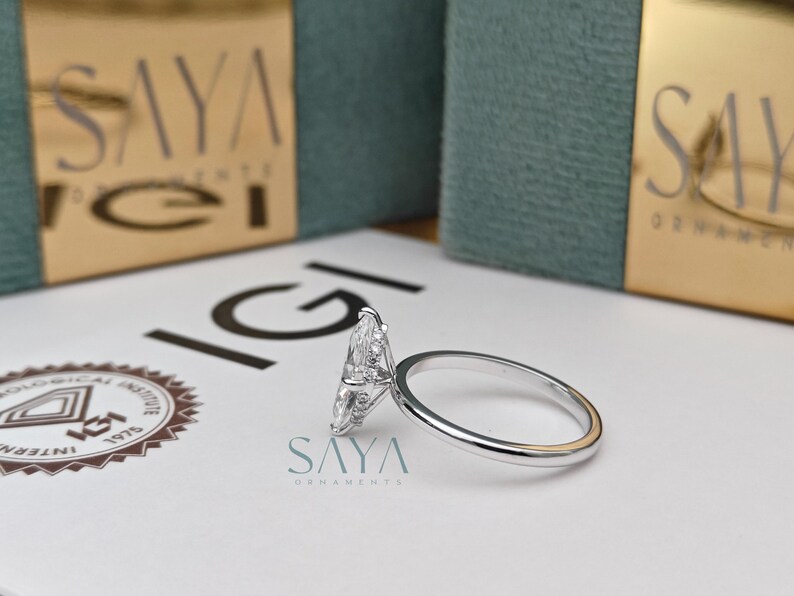 Puede incluir: Un anillo de plata con un diamante talla marquesa, sobre una superficie blanca. El anillo est&aacute; junto a una caja con el texto "SAYA ORNAMENTS". Es una joya.