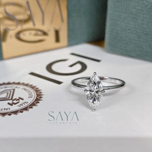 Puede incluir: Un anillo de diamantes con talla marquesa y una banda plateada. El anillo est&aacute; centrado en una superficie blanca con el texto "IGI" y "SAYA" visible. El fondo incluye cajas con detalles dorados y verde azulado.