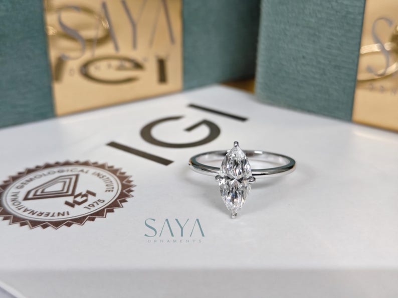 Puede incluir: Un anillo de diamantes con talla marquesa y una banda plateada. El anillo est&aacute; centrado en una superficie blanca con el texto "IGI" y "SAYA" visible. El fondo incluye cajas con detalles dorados y verde azulado.