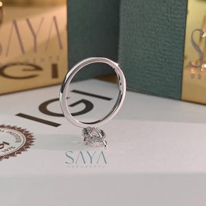 Puede incluir: Un anillo de plata con un diamante en forma de pera. El anillo se muestra sobre una superficie blanca, con el texto "SAYA ORNAMENTS" visible en el fondo. El anillo es el punto focal de la imagen.