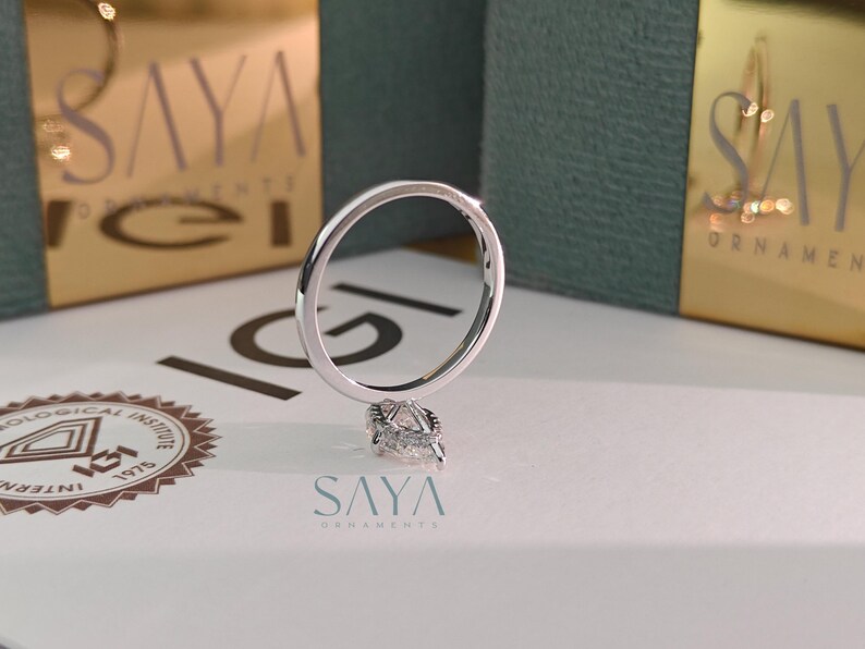 Puede incluir: Un anillo de plata con un diamante en forma de pera. El anillo se muestra sobre una superficie blanca, con el texto "SAYA ORNAMENTS" visible en el fondo. El anillo es el punto focal de la imagen.