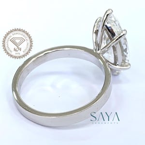 May include: A silver engagement ring with a large, pear-shaped diamond. The diamond is held in place by four prongs. The ring band is smooth and polished. The image also includes the text "SAYA ORNAMENTS" and a logo.