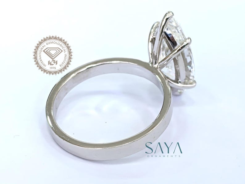 May include: A silver engagement ring with a large, pear-shaped diamond. The diamond is held in place by four prongs. The ring band is smooth and polished. The image also includes the text "SAYA ORNAMENTS" and a logo.