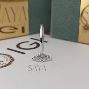 Puede incluir: Un anillo de plata con un diamante central de talla marquesa, rodeado de diamantes m&aacute;s peque&ntilde;os. El anillo est&aacute; sobre una superficie blanca con el texto "SAYA ORNAMENTS". El fondo incluye embalajes dorados y turquesa.