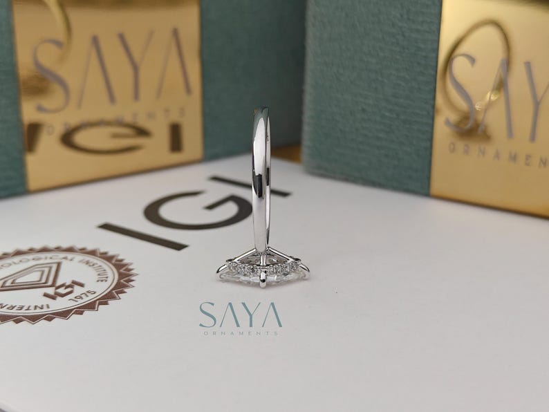 Puede incluir: Un anillo de plata con un diamante central de talla marquesa, rodeado de diamantes m&aacute;s peque&ntilde;os. El anillo est&aacute; sobre una superficie blanca con el texto "SAYA ORNAMENTS". El fondo incluye embalajes dorados y turquesa.