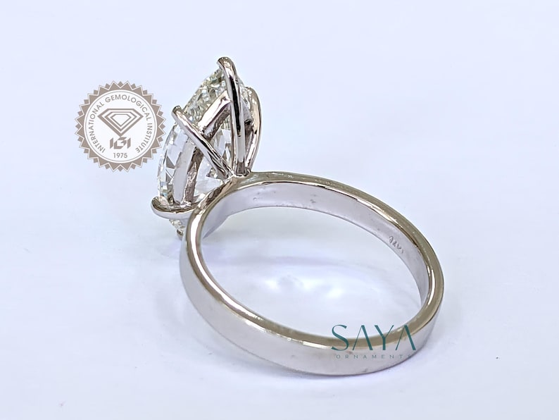 May include: A close-up of a platinum ring with a large, pear-shaped diamond. The ring is set with prongs and features a smooth, polished band. The image includes the text "SAYA ORNAMENT" and a logo from the International Gemological Institute.