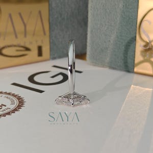 Puede incluir: Primer plano de un anillo plateado con un dise&ntilde;o geom&eacute;trico. El anillo est&aacute; sobre una superficie blanca con el logotipo "SAYA" en color turquesa. El fondo incluye cajas doradas con el nombre y el logotipo de la marca.