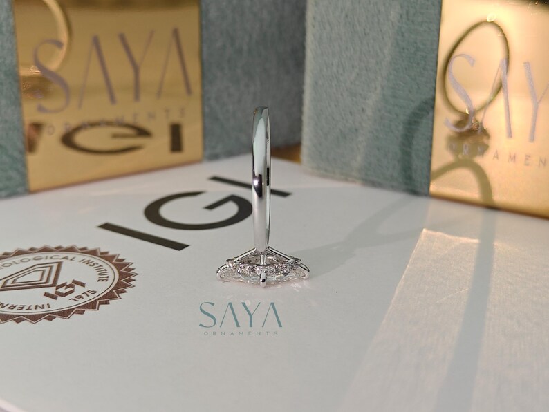 Puede incluir: Primer plano de un anillo plateado con un dise&ntilde;o geom&eacute;trico. El anillo est&aacute; sobre una superficie blanca con el logotipo "SAYA" en color turquesa. El fondo incluye cajas doradas con el nombre y el logotipo de la marca.