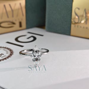Puede incluir: Un anillo de diamantes con talla marquesa y banda plateada sobre una superficie blanca. El anillo, con su forma alargada y puntiaguda, es el foco. El fondo incluye cajas con letras doradas y el logo "SAYA" y IGI.