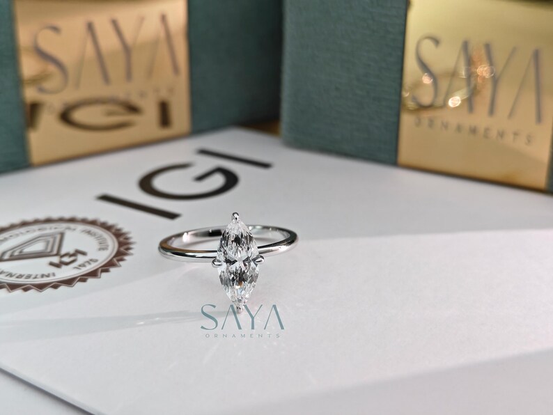 Puede incluir: Un anillo de diamantes con talla marquesa y banda plateada sobre una superficie blanca. El anillo, con su forma alargada y puntiaguda, es el foco. El fondo incluye cajas con letras doradas y el logo "SAYA" y IGI.