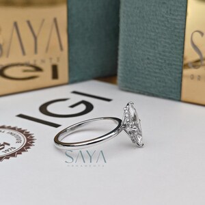Puede incluir: Un anillo de diamantes con corte marquesa y banda plateada descansa sobre una superficie blanca. El anillo est&aacute; colocado cerca de un logotipo y el texto "SAYA ORNAMENTS". El fondo incluye dos cajas turquesas con letras doradas.