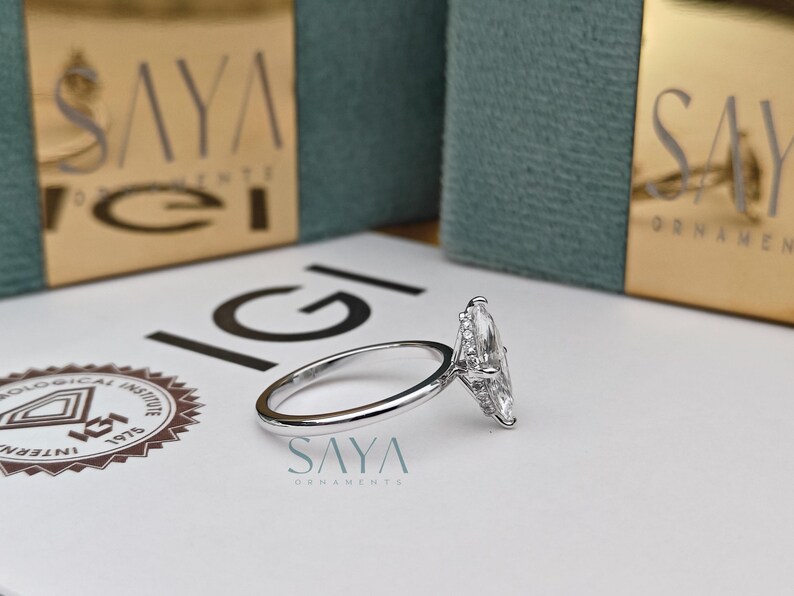 Puede incluir: Un anillo de diamantes con corte marquesa y banda plateada descansa sobre una superficie blanca. El anillo est&aacute; colocado cerca de un logotipo y el texto "SAYA ORNAMENTS". El fondo incluye dos cajas turquesas con letras doradas.