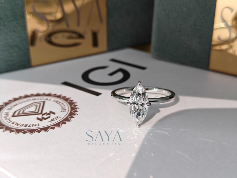 Puede incluir: Un anillo de diamantes con talla marquesa y una fina banda plateada. El diamante es el punto focal, reflejando la luz. El anillo est&aacute; sobre una superficie blanca con el texto "SAYA" y un logotipo. El fondo incluye una caja.