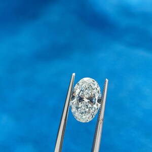 May include: A clear oval-shaped diamond held by silver tweezers against a blue background.
