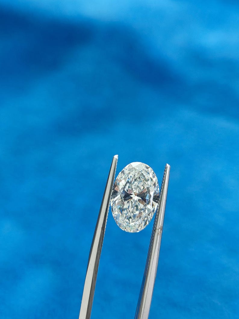 May include: A clear oval-shaped diamond held by silver tweezers against a blue background.