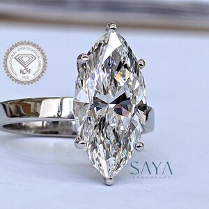 May include: A marquise-cut diamond engagement ring with a polished silver band. The large, clear diamond is the focal point, reflecting light. The ring is set against a soft background. The ring is from Saya Ornaments.