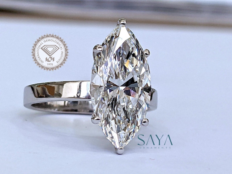 May include: A marquise-cut diamond engagement ring with a polished silver band. The large, clear diamond is the focal point, reflecting light. The ring is set against a soft background. The ring is from Saya Ornaments.