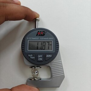 May include: A digital caliper measuring a clear gemstone. The caliper reads 11.87 mm. The caliper has a black display with white numbers and a silver body. The caliper has buttons labeled "inch/mm", "OFF/ON", and "ZERO".