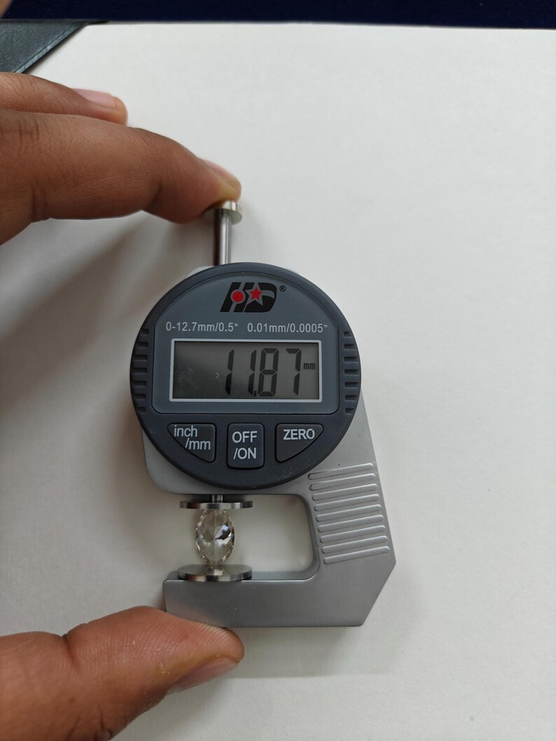 May include: A digital caliper measuring a clear gemstone. The caliper reads 11.87 mm. The caliper has a black display with white numbers and a silver body. The caliper has buttons labeled "inch/mm", "OFF/ON", and "ZERO".