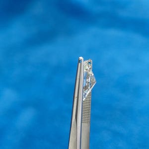May include: A clear, faceted diamond gemstone held by silver tweezers against a blue background.