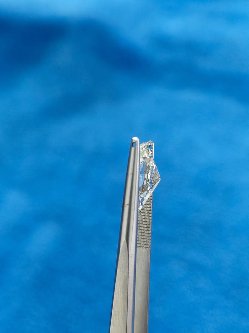 May include: A clear, faceted diamond gemstone held by silver tweezers against a blue background.