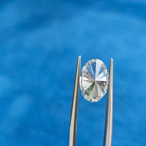 May include: A clear oval-shaped diamond held by tweezers against a blue background.