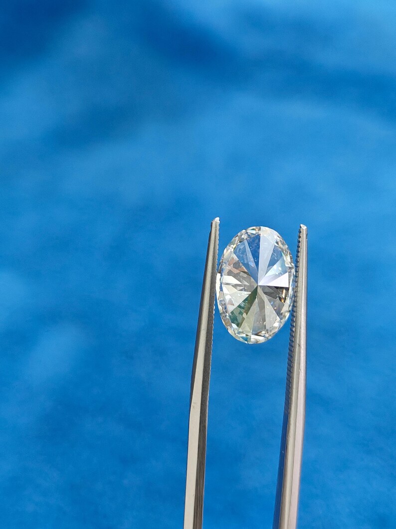 May include: A clear oval-shaped diamond held by tweezers against a blue background.
