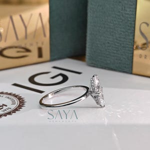 Puede incluir: Un anillo de plata con una gran piedra central de diamante en forma de pera y piedras de acento m&aacute;s peque&ntilde;as. El anillo est&aacute; sobre una superficie blanca con el nombre de la marca "SAYA" visible.