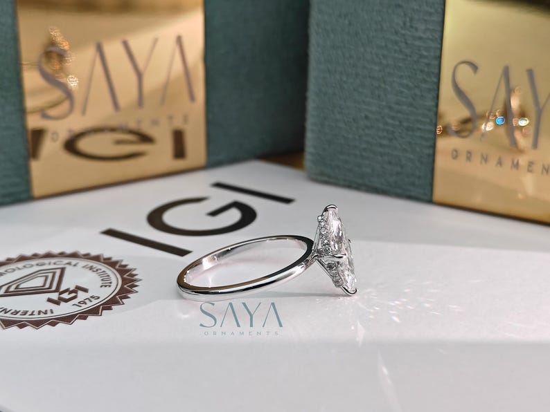 Puede incluir: Un anillo de plata con una gran piedra central de diamante en forma de pera y piedras de acento m&aacute;s peque&ntilde;as. El anillo est&aacute; sobre una superficie blanca con el nombre de la marca "SAYA" visible.
