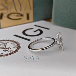 Puede incluir: Un anillo de compromiso plateado con un diamante talla marquesa. El anillo est&aacute; sobre una superficie blanca con el logo IGI y el texto "SAYA ORNAMENTS". El fondo incluye una caja dorada y verde.