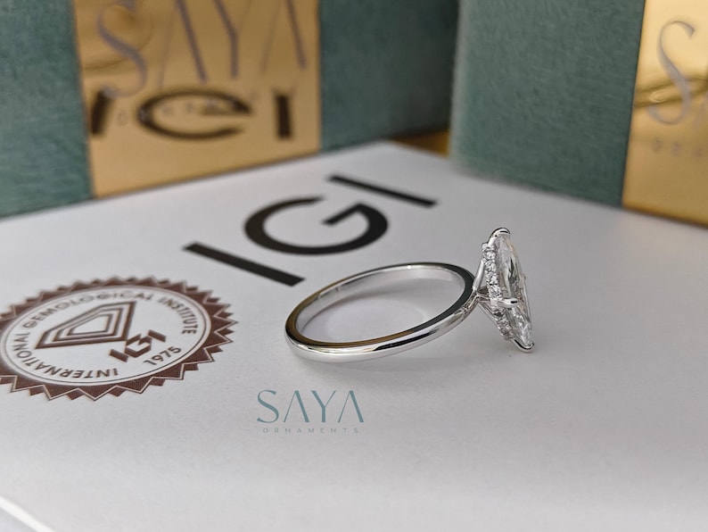 Puede incluir: Un anillo de compromiso plateado con un diamante talla marquesa. El anillo est&aacute; sobre una superficie blanca con el logo IGI y el texto "SAYA ORNAMENTS". El fondo incluye una caja dorada y verde.
