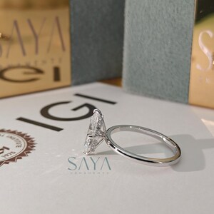 Puede incluir: Un anillo de compromiso de diamantes con una piedra central en forma de pera y una fina banda plateada. El anillo est&aacute; sobre una superficie blanca con el texto "IGI" y "SAYA ORNAMENTS". Una caja dorada est&aacute; en el fondo.