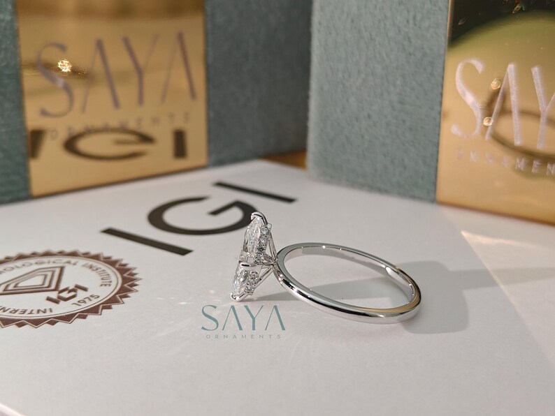 Puede incluir: Un anillo de compromiso de diamantes con una piedra central en forma de pera y una fina banda plateada. El anillo est&aacute; sobre una superficie blanca con el texto "IGI" y "SAYA ORNAMENTS". Una caja dorada est&aacute; en el fondo.