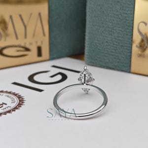 Puede incluir: Un anillo de plata con un diamante en forma de pera. El anillo est&aacute; sobre una superficie blanca, con el logotipo de IGI y el logotipo de Saya en el fondo. Una joya.