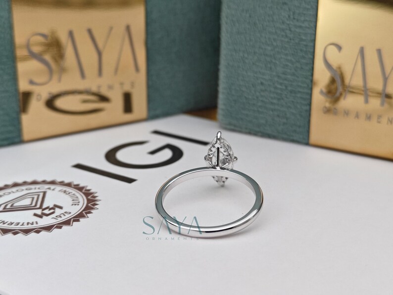 Puede incluir: Un anillo de plata con un diamante en forma de pera. El anillo est&aacute; sobre una superficie blanca, con el logotipo de IGI y el logotipo de Saya en el fondo. Una joya.