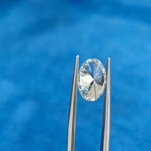 May include: A clear, round, faceted gemstone held by tweezers against a blue background.