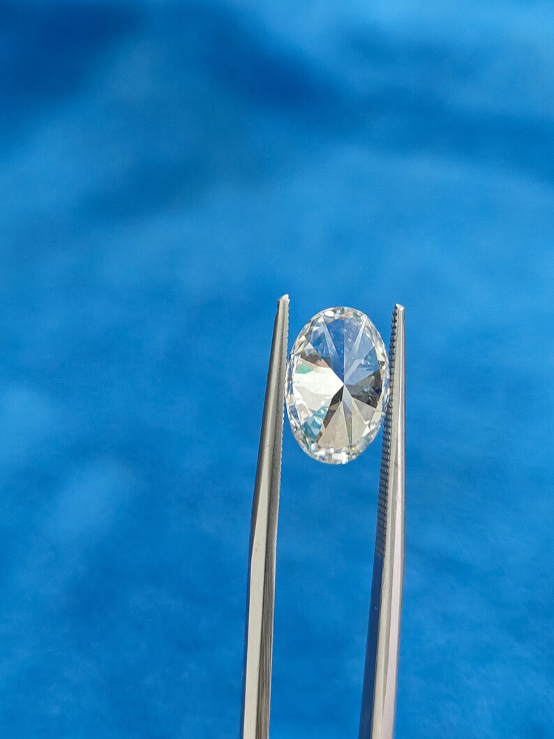 May include: A clear, round, faceted gemstone held by tweezers against a blue background.