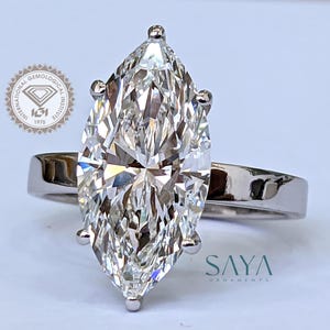 May include: A marquise-cut diamond ring with a polished silver band. The large, clear diamond is the focal point, set with four prongs. The ring is displayed on a white surface. The logo of the International Gemological Institute is visible.