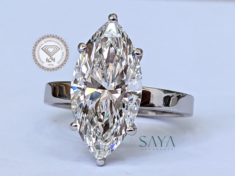May include: A marquise-cut diamond ring with a polished silver band. The large, clear diamond is the focal point, set with four prongs. The ring is displayed on a white surface. The logo of the International Gemological Institute is visible.