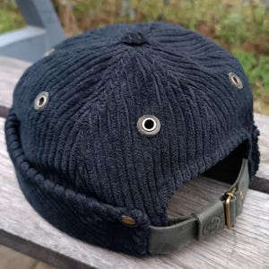 Könnte beinhalten: Eine schwarze Cord-Newsboy-Mütze mit einem Lederriemen und Metallösen. Die Mütze hat eine abgerundete Krone und ein Schild, das nach oben oder unten geklappt werden kann.