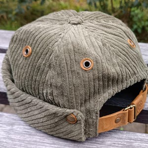 May include: A green corduroy bucket hat with a brown leather strap and metal eyelets. The hat is sitting on a wooden surface.