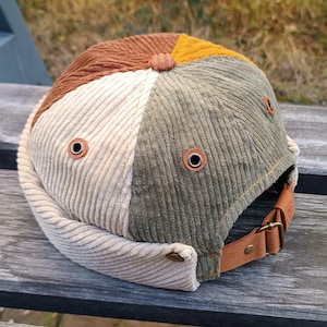 May include: A corduroy baseball cap with a patchwork design in beige, brown, green, and yellow. The cap has a leather strap with a metal buckle.
