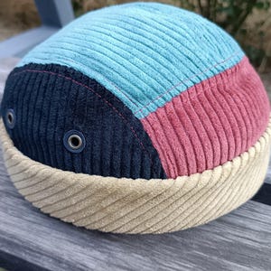 May include: A colorful corduroy hat with a unique design. The hat features sections of light blue, red, and navy blue corduroy, with a tan corduroy brim. Two metal eyelets are visible on the navy section.