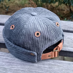 May include: A grey corduroy cap with a short brim and a brown leather strap. The cap has a rounded crown and features several metal eyelets with brown leather reinforcements. The adjustable strap fastens with a metal buckle.