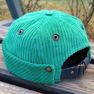 Könnte beinhalten: Eine grüne Cord-Newsboy-Mütze mit einem schwarzen Lederriemen und Messing-Akzenten.