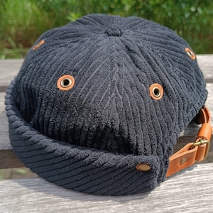 Peut inclure: Casquette en velours côtelé noir avec une couronne arrondie et un petit bord. La casquette comporte plusieurs œillets métalliques avec des accents en cuir marron et une sangle en cuir marron avec une boucle en métal.