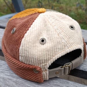 May include: A brown, beige, and yellow corduroy beanie with a leather strap and metal eyelets.