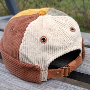 Può includere: Cappellino in velluto a coste con un design patchwork nei toni del marrone, beige, giallo e verde. Il cappellino presenta un cinturino in pelle marrone con fibbia in metallo e occhielli in metallo. Un accessorio alla moda.