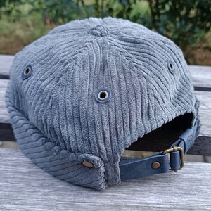 May include: A grey corduroy cap with a short peak and a leather strap. The cap features a classic design with metal eyelets and a brass buckle. The fabric has a ribbed texture, and the cap is likely adjustable for a comfortable fit.